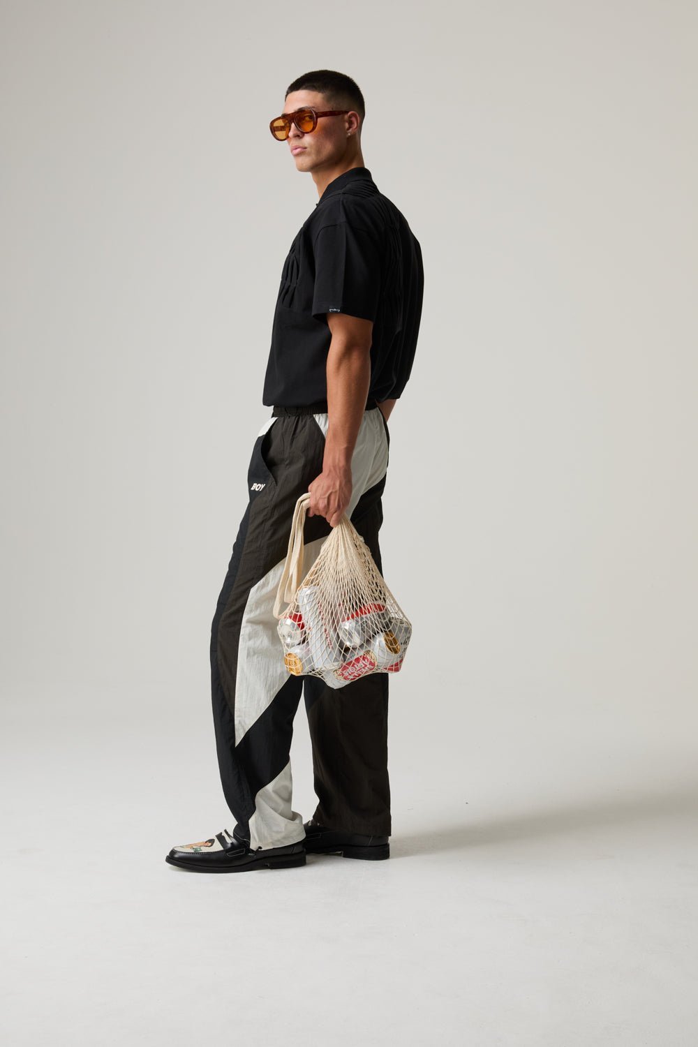 Man wearing black shirt and patterned pants holding a mesh shopping bag, standing against white background